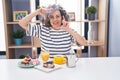 Middle age woman with grey hair eating pastries and drinking coffee for breakfast smiling making frame with hands and fingers with
