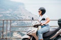 Middle-age woman in helmet and sunglasses on motor scooter on the Sicilian old town streets in the Forza d`Agro with Sant`Alessi