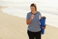 Middle age woman holding a sports mat and preparing to practice yoga outdoors on sea beach. Happy mature overweight Royalty Free Stock Photo