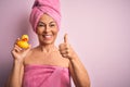 Middle age woman wearing pink bath towel from beauty body care holding rubber yellow duck happy with big smile doing ok sign,