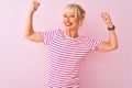 Middle age woman wearing striped t-shirt standing over isolated pink background showing arms muscles smiling proud