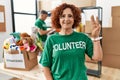 Middle age woman wearing volunteer t shirt at donations stand showing and pointing up with fingers number three while smiling
