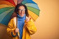 Middle age woman wearing yellow raincoat under colorful umbrella over isolated background pointing and showing with thumb up to Royalty Free Stock Photo