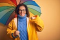 Middle age woman wearing yellow raincoat under colorful umbrella over isolated background with surprise face pointing finger to Royalty Free Stock Photo
