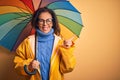 Middle age woman wearing yellow raincoat under colorful umbrella over isolated background very happy pointing with hand and finger Royalty Free Stock Photo