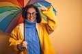 Middle age woman wearing yellow raincoat under colorful umbrella over isolated background very happy pointing with hand and finger Royalty Free Stock Photo