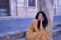 Middle-aged adult Hispanic woman with black curly hair, wearing a colorful outfit, sitting on stone steps, pensive. Concept Royalty Free Stock Photo