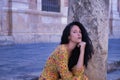 Middle-aged adult Hispanic woman with black curly hair, wearing a colorful outfit, sitting on stone steps, pensive. Concept Royalty Free Stock Photo