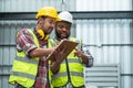 Middle aged African engineer and Asian foreman in safety hat working together at warehouse while listing on clipboard to discuss, Royalty Free Stock Photo