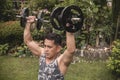 A middle aged asian man in a camouflage patterned tank top does standing dumbbell shoulder presses at his front yard.