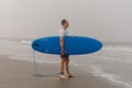 Mature athlete keeping blue surfboard under armpit standing on wet sandy shore in front of sea and looking at seascape.