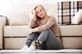 Middle aged barefoot woman sitting at the floor embracing her knees, near sofa at home, her head down, bored, troubled Royalty Free Stock Photo