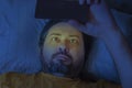 A middle-aged bearded man lies on a pillow late at night and looks at the screen of a mobile phone. Royalty Free Stock Photo