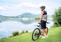 A middle-aged bicyclist man dressed in cycling clothes, helmet, and sunglasses with bicycle on grass hill enjoying lake and