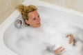 Middle-aged blonde woman relaxes immersed in the bathtub filled with water and foam