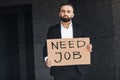 Middle-aged businessman in suit holding sign with words NEED JOB while standing near office building, free space Royalty Free Stock Photo