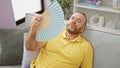 Middle-aged caucasian man suffering in hot weather, fanning himself with handfan while sitting on sofa at home