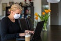 Middle aged Caucasian woman working from home on laptop in her living room, wearing face protective mask whilst being in Royalty Free Stock Photo
