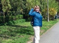 Middle-aged European woman, blonde, cheerfully smiles and waves her hand, stands in a park with flowering trees