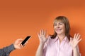 Middle-aged European woman stands protestingly extending both hands, male hand holds the phone, orange background, close-up, copy