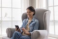 Middle-aged female sit rest on cozy armchair with modern cellphone