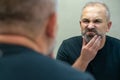 Middle-aged handsome man looking in mirror in bathroom touching his beard thinking of cutting it off