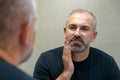 Middle-aged handsome man looking in mirror in bathroom touching his beard thinking of cutting it off