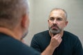 Middle-aged handsome man looking in mirror in bathroom touching his beard thinking of cutting it off