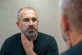 Middle-aged handsome man looking in mirror in bathroom touching his beard thinking of cutting it off