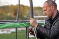 Middle aged human with furrowed eyebrows carefully fastening metal posts to trampoline support outdoor using bolt. Royalty Free Stock Photo