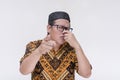 A middle aged indonesian man covers his nose while pointing at someone, complaining at their body odor. Wearing a batik shirt