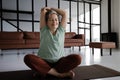 A middle aged lady practices yoga at home. A smiling woman with closed eyes in bliss sits on a mat on the floor in the