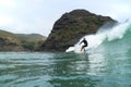 A middle aged male surfer in a black wetsuit rides a wave with cliffs, coastal hills and a blue cloudy sky in the background at