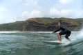 A middle aged male surfer in a black wetsuit rides a wave with cliffs, coastal hills and a blue cloudy sky in the background at