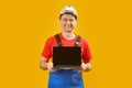 Middle aged man architect engineer wears safety helmet which hand holding laptop with empty screen on yellow studio background. Royalty Free Stock Photo