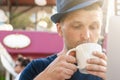 A middle-aged man in a blue hat is drinking coffee in front of a laptop. portrait close-up, looking into a mug. vertical photo