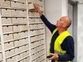 A middle-aged man in a blue uniform and yellow vest. A worker pulls out wooden boxes in a large storage system signed with numbers