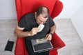 Middle-aged man in glasses with a laptop on his lap sitting in a red chair and counting money Royalty Free Stock Photo