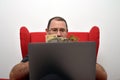 Middle-aged man in glasses with a laptop on his lap sitting in a red chair and counting money Royalty Free Stock Photo