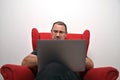 Middle-aged man in glasses with a laptop on his lap is sitting in a red armchair, front view Royalty Free Stock Photo