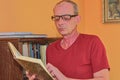 Middle aged man man is reading book in living room. Mature man is standing next to bookcase