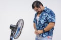 Middle aged man seeking relief from the heat by unbuttoning his Hawaiian shirt in front of an electric fan, isolated on white Royalty Free Stock Photo