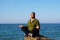 Middle aged man sits in lotus position on a large rock by the sea and meditates in sunlight, solitude in nature