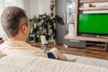 A middle-aged man sits on a sofa in the living room and watches TV with a remote control