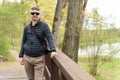 A middle-aged man in sunglasses and a thin down jacket on a wooden footbridge near the lake in early spring