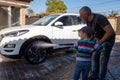 A middle-aged man teaches children of boys 4 and 10 years old to wash a car in the yard of his house on a summer sunny day. 2019. Royalty Free Stock Photo