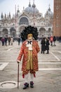 A middle-aged man in a vintage costume with a lace collar, wearing a carnival mask bordered by a circle of high black feathers,