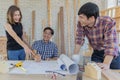 Middle aged man wearing blue plaid shirt holding pencil explaining and discussing blueprint document to young male and female Royalty Free Stock Photo