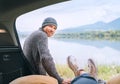 A middle-aged man dressed in warm knitted clothes near the car smiling at a woman in a car trunk who enjoying a mountain lake view