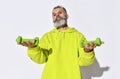 Middle aged positive brutal bearded man in bright yellow green casual hoodie standing and showing green dumbbels